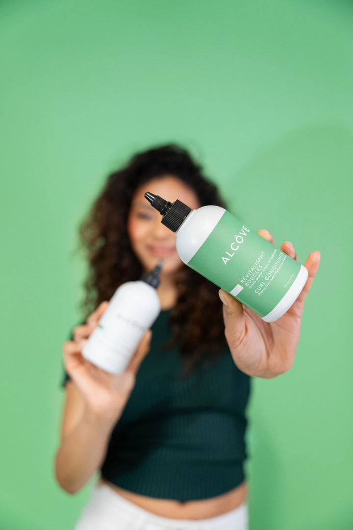 Femme souriante tenant le revitalisant boucles Alcôve au premier plan, sur fond vert, mettant en avant le produit pour cheveux texturés.