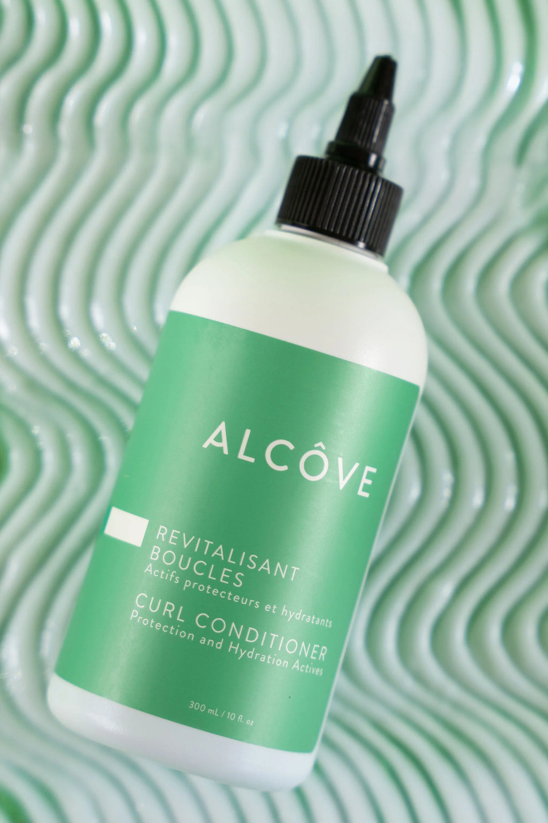 Flacon de revitalisant boucles Alcôve en gros plan, posé sur un fond vert texturé en forme de vagues, mettant en valeur le design du produit.