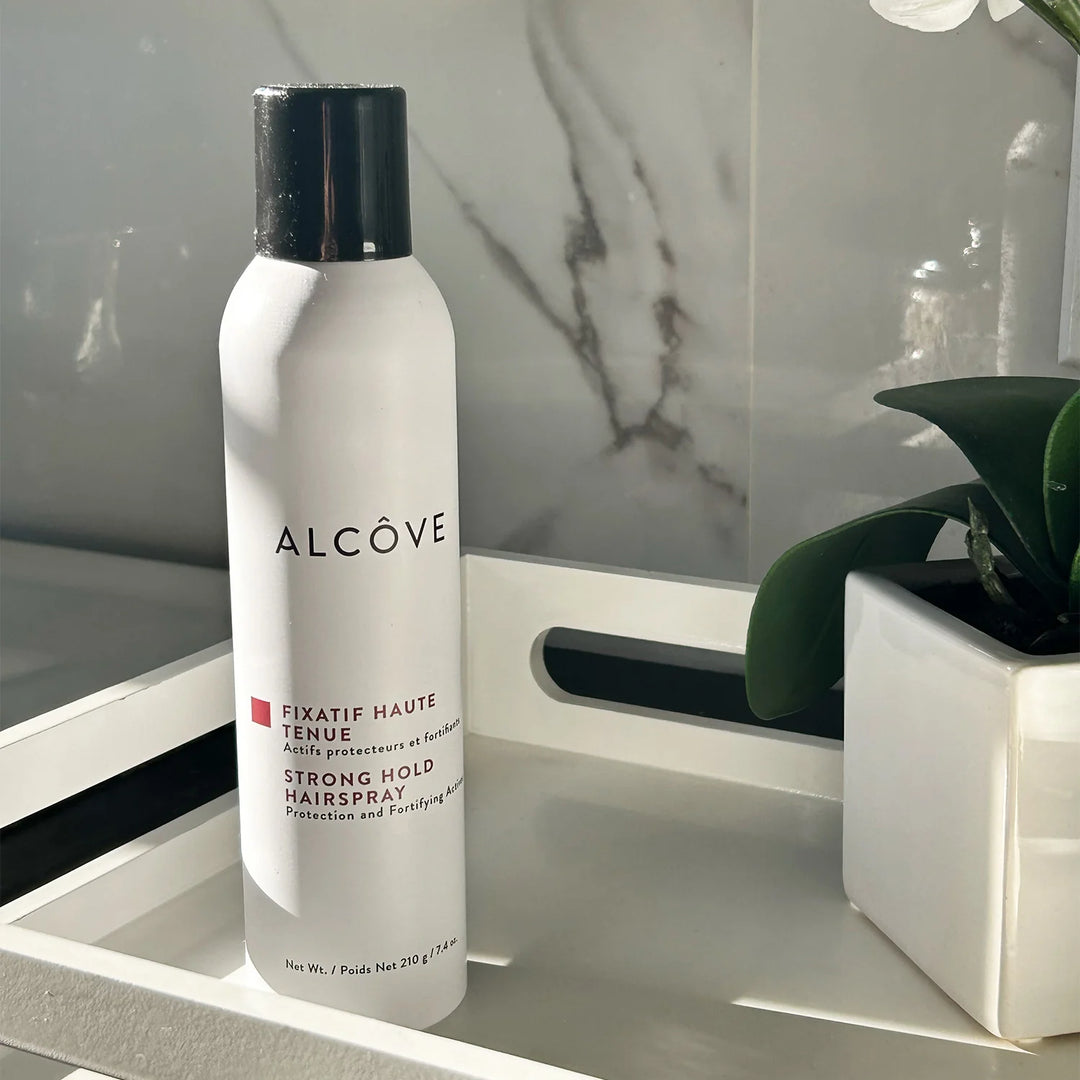 Spray coiffant Alcôve haute tenue de 210 g posé sur un plateau blanc dans une salle de bain moderne avec mur en marbre et plante décorative — fixatif cheveux avec protection solaire UV, idéal pour prévenir les frisottis et prolonger la couleur.
