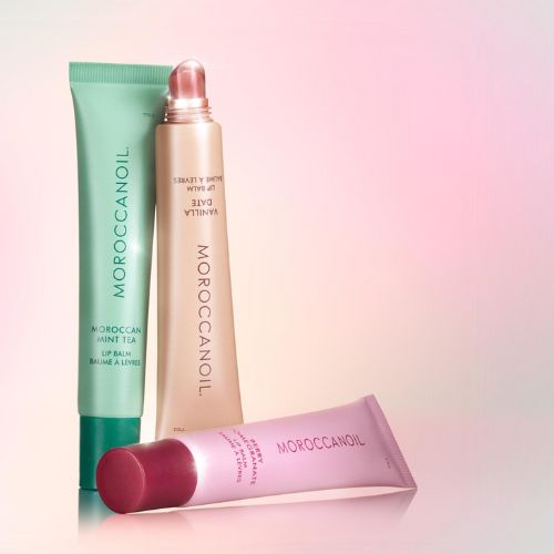 Baumes à lèvres Moroccanoil – tubes Mint Tea, Vanilla Date et Berry Pomegranate posés sur fond dégradé rose, effet lumineux et packaging élégant.