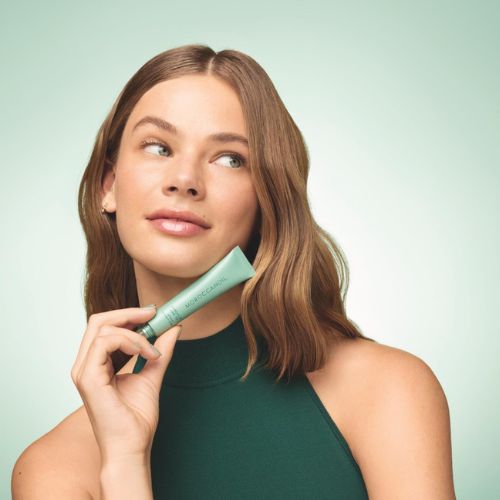 Femme souriante tenant le baume à lèvres Moroccanoil Mint Tea – ambiance fraîche et naturelle, fond vert doux, style beauté minimaliste.