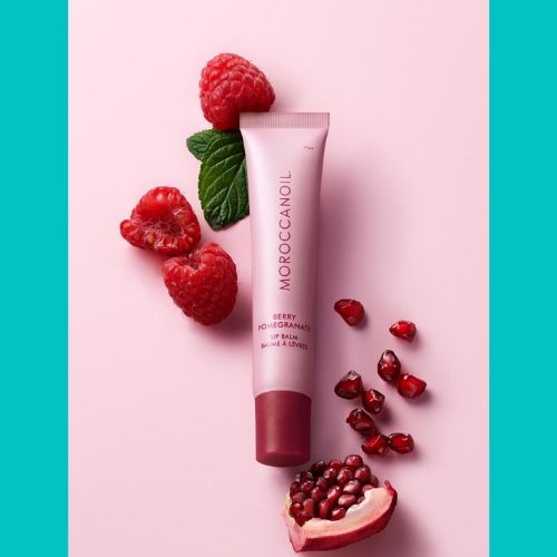 Baume à lèvres Moroccanoil Berry Pomegranate – tube rose 15g sur fond rose avec framboises fraîches, graines de grenade et feuilles de menthe.