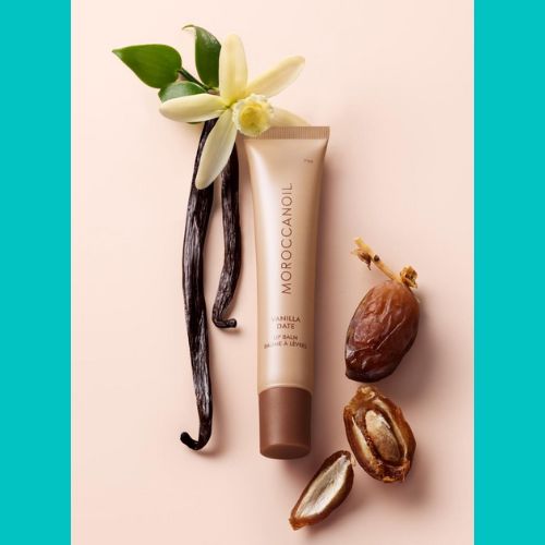 Baume à lèvres Moroccanoil Vanilla Date – tube beige doré sur fond nude avec gousses de vanille, fleurs de vanille et dattes ouvertes.