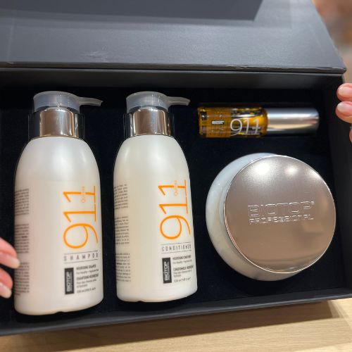Coffret ouvert Biotop 911 contenant un shampoing 330ml, un revitalisant 330ml, un masque 350ml et un sérum 30ml. Produits capillaires pour cheveux secs et endommagés, présentés dans un écrin noir raffiné avec mains tenant le couvercle, sur fond de salon professionnel.