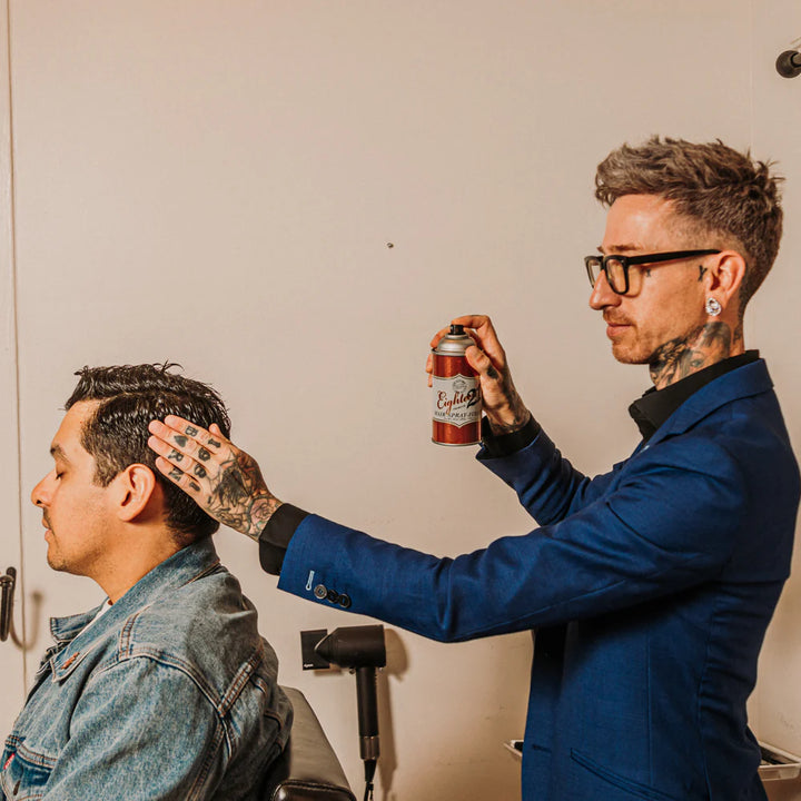 Coiffeur tatoué en veste bleue utilisant la laque coiffante 18.21 Man Made sur un client assis en salon, cheveux courts coiffés, dans un cadre professionnel. Aérosol au design rétro rouge et blanc avec étiquette visible. Scène en intérieur, éclairage chaud, ambiance de barbier moderne.