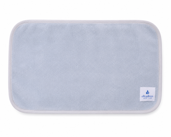 Serviette nettoyante pour le visage Eltraderm en microfibres gris pâle avec bordure assortie et étiquette de marque visible, présentée à plat sur fond blanc. Accessoire réutilisable et lavable conçu pour nettoyer la peau en douceur.
