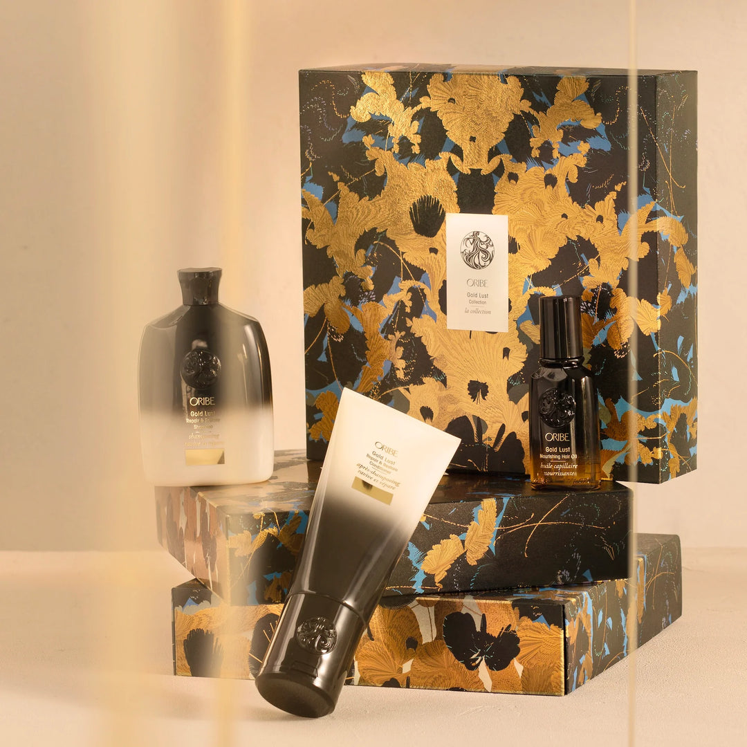 Coffret Oribe Gold Lust en édition luxueuse, décor floral noir, doré et bleu, accompagné de trois produits capillaires : shampooing, revitalisant et huile capillaire. Présentation élégante sur fond doux et lumineux, ambiance raffinée.
