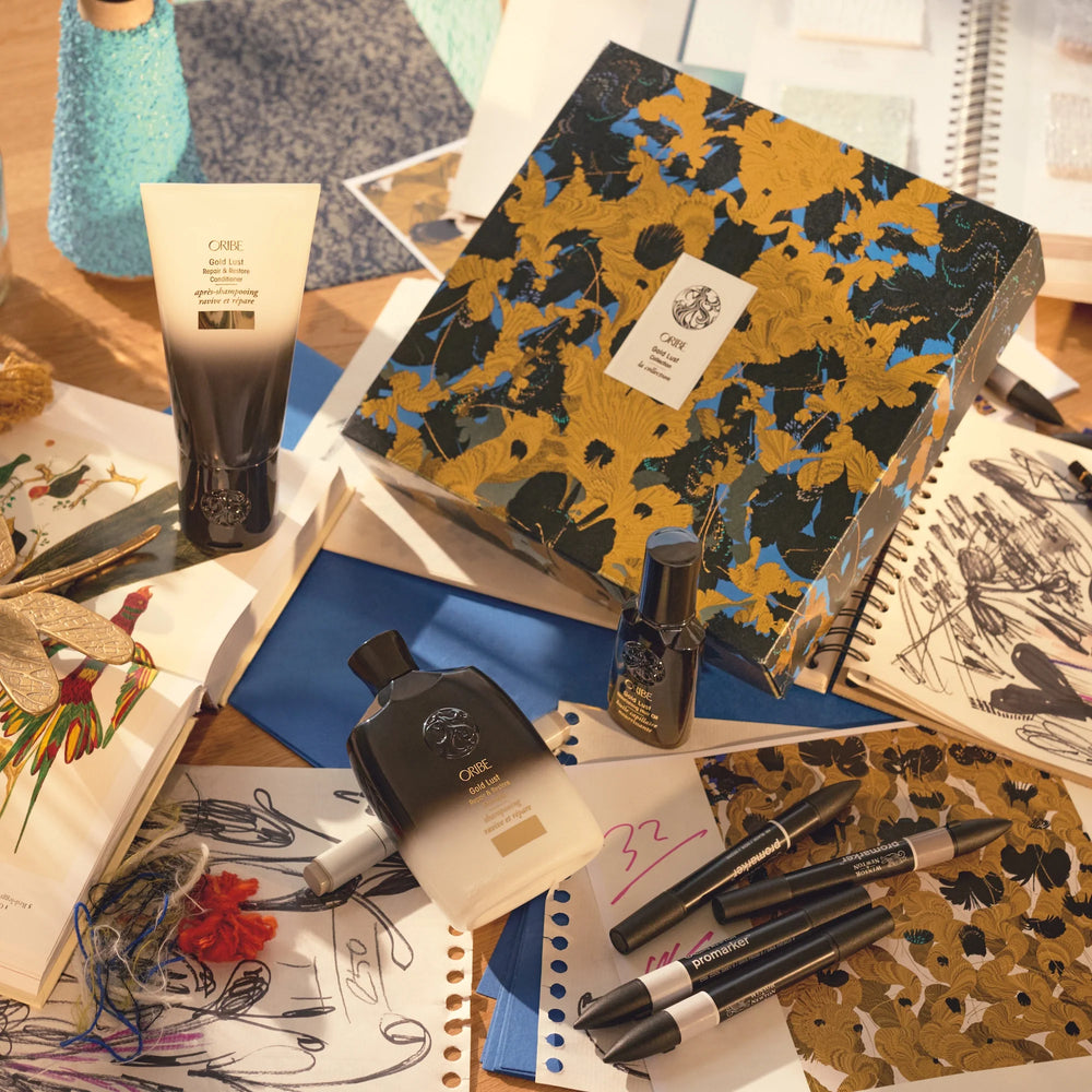 Coffret Oribe Gold Lust sur un bureau créatif, entouré de dessins, carnets et feutres. Les trois soins cheveux – shampooing, revitalisant et huile – sont dispersés autour de la boîte décorative noire à motifs dorés et bleus. Ambiance artistique et haut de gamme.