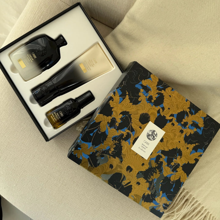 Coffret de soins capillaires Oribe Gold Lust ouvert, présenté sur tissu beige. Trois produits alignés dans l’écrin : shampooing, revitalisant et huile en flacons élégants, dans une boîte décorative noir, or et bleu.