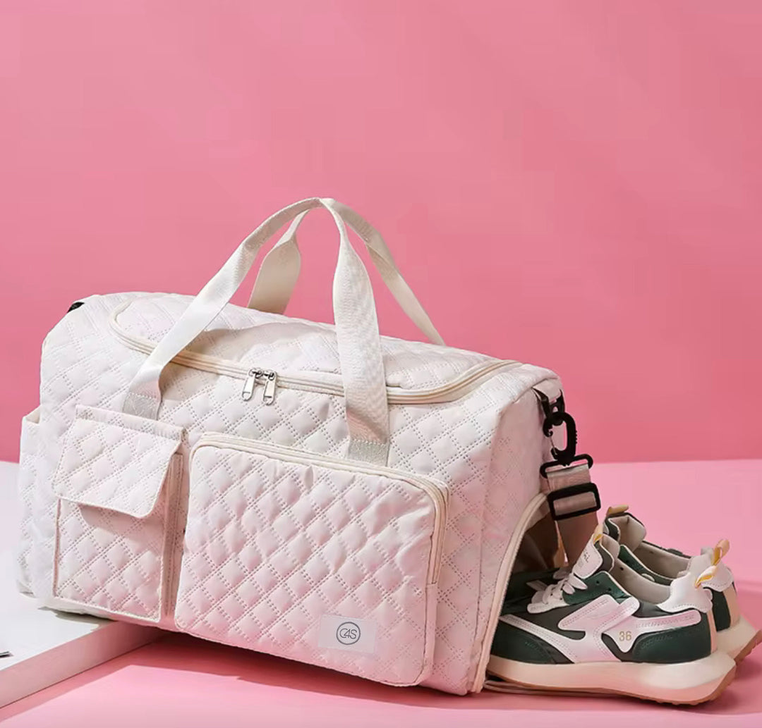 Sac de sport matelassé crème C4S déposé sur un fond rose. Le sac comporte des poignées beiges, une grande poche avant à fermeture éclair avec une étiquette cousue C4S bleu pâle, et un compartiment latéral où sortent une paire d’espadrilles vertes et blanches. L’image évoque un style chic, doux et fonctionnel.