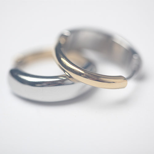 Paire de boucles d’oreilles créoles épaisses, une en argent poli et l’autre en finition bicolore argent et or jaune, posées entrelacées sur fond blanc. Design arrondi, minimaliste et moderne.
