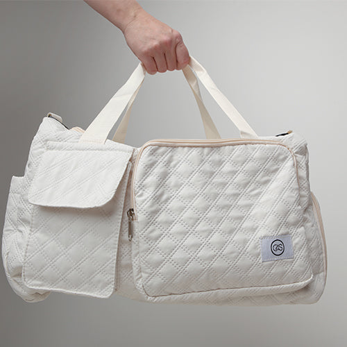 Sac de voyage matelassé blanc cassé avec poignées en toile écrue, grandes poches extérieures zippées et à rabat, porté à la main sur fond neutre. Design pratique, léger et élégant.