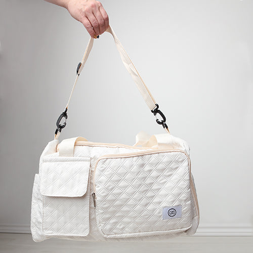 Sac de voyage matelassé blanc cassé avec bandoulière réglable détachable de couleur écrue, porté à la main. Poches extérieures zippées et à rabat, design élégant et pratique, présenté sur fond blanc.