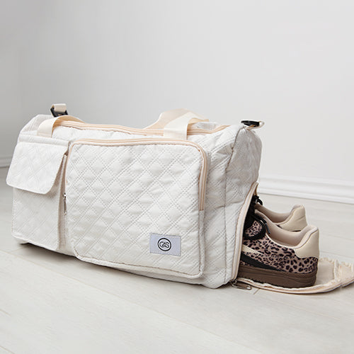 Sac de voyage matelassé blanc cassé avec grandes poches extérieures et poignées écrues, posé au sol sur parquet clair. Compartiment latéral zippé ouvert laissant apparaître une paire de chaussures de sport beige à motif léopard. Design fonctionnel et élégant.
