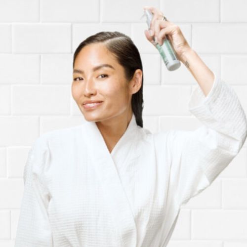 Femme souriante en peignoir blanc appliquant l’essence Joico Scalp Vitality directement sur son cuir chevelu à l’aide du spray, dans une salle de bain carrelée blanche.