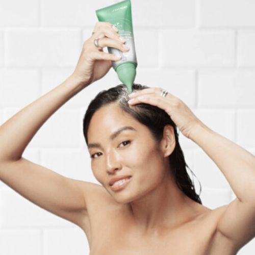 Femme aux cheveux mouillés appliquant le gommage Joico Scalp Vitality directement sur le cuir chevelu, en massant doucement avec les doigts dans une salle de bain blanch