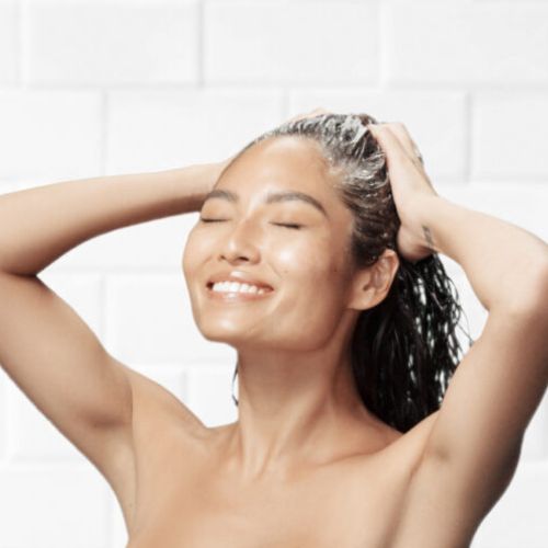 Femme souriante aux cheveux mouillés massant son cuir chevelu sous la douche, illustrant l'utilisation du shampooing Joico Scalp Vitality dans un moment de bien-être.