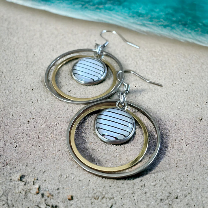 Paire de boucles d’oreilles en acier inoxydable posée sur le sable, avec anneaux doré et argenté, et cabochon central rond à rayures noires et blanches.