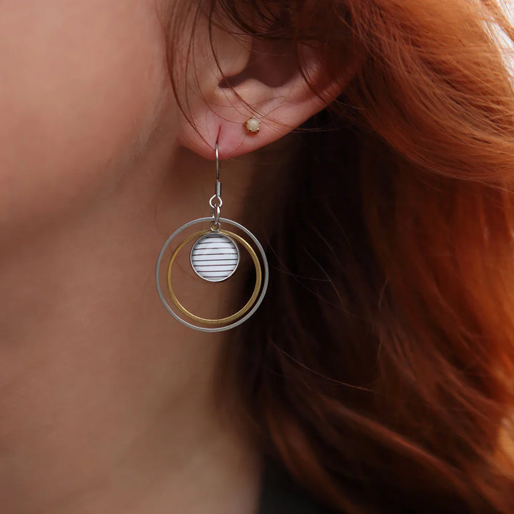 Boucle d’oreille pendante portée à l’oreille, composée de deux anneaux métalliques doré et argenté, et d’un cabochon rayé noir et blanc.