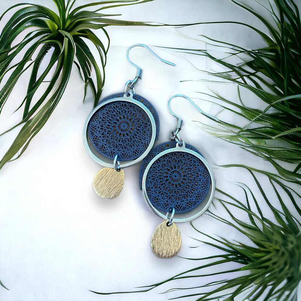 Paire de boucles d’oreilles bleu marine avec cercle ajouré, anneau argenté et petite goutte dorée, posées à plat sur fond blanc entourées de feuillage.
