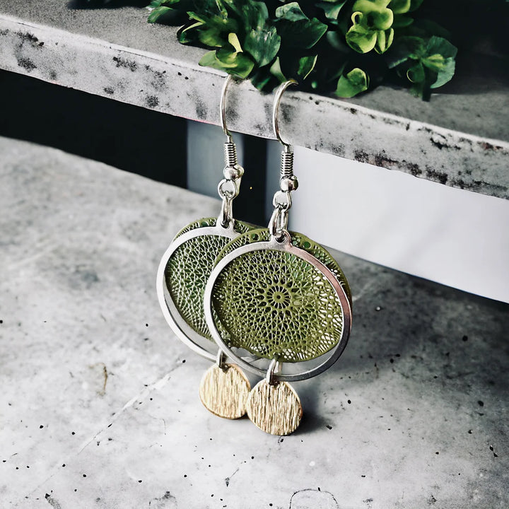 Boucles d’oreilles pendantes vert kaki et dorées, accrochées à une surface grise. Elles comportent un cercle texturé, un anneau argenté et un pendentif goutte en laiton brossé.