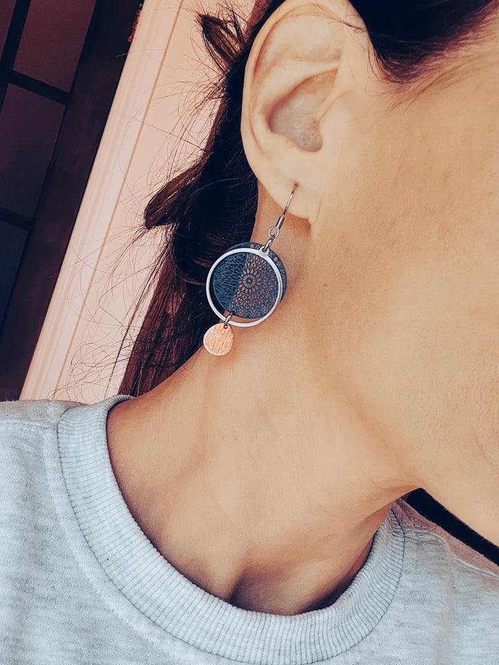 Vue rapprochée d’une femme portant une boucle d’oreille pendante bleu marine avec cercle texturé, anneau argenté et goutte dorée brossée, mise en valeur par une tenue décontractée.