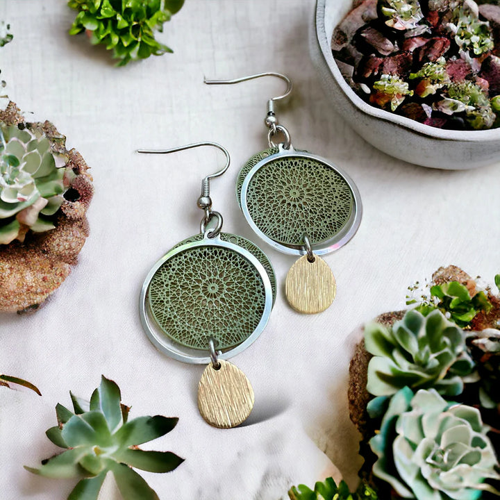 Paire de boucles d’oreilles artisanales vert kaki avec motif mandala, anneau argenté et petite goutte dorée, posées sur une nappe blanche entourée de plantes grasses.