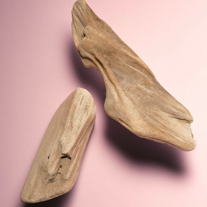 Morceaux de bois naturel clair avec texture organique posés sur un fond rose, évoquant des notes boisées chaudes utilisées en parfumerie et en soins.