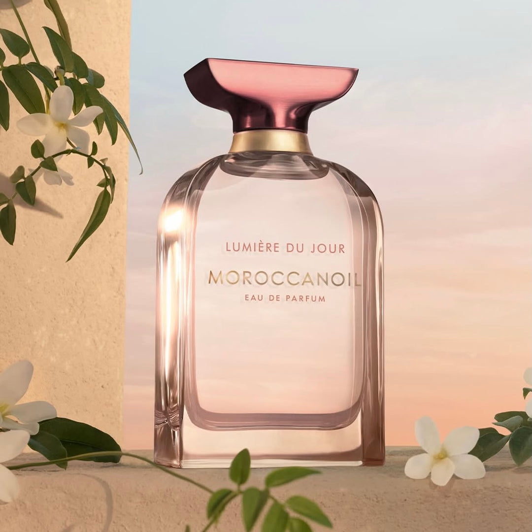 Flacon de parfum Moroccanoil Lumière du Jour eau de parfum posé dans un décor lumineux avec fleurs blanches et ciel au coucher du soleil.