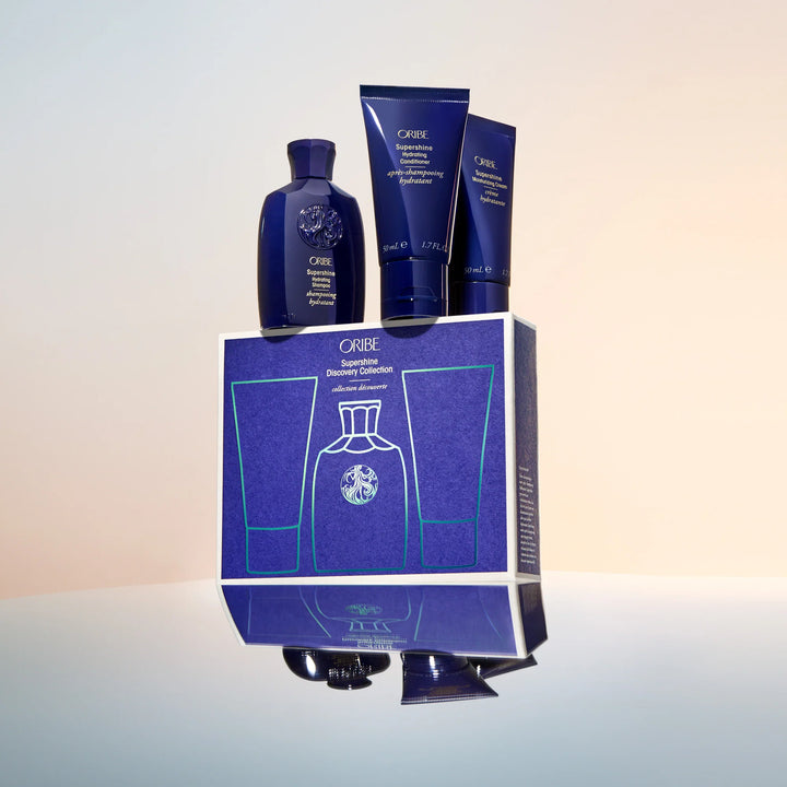 Coffret Oribe Supershine Discovery Collection avec shampooing, après-shampooing et crème hydratante formats voyage disposés sur la boîte du coffret au design bleu signature.