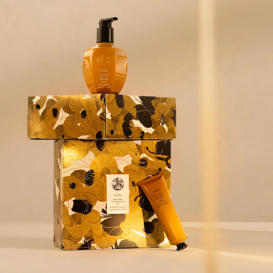 Flacon de savon pour les mains Oribe Côte d’Azur posé sur un coffret doré fleuri, avec la crème pour les mains assortie au pied du coffret.