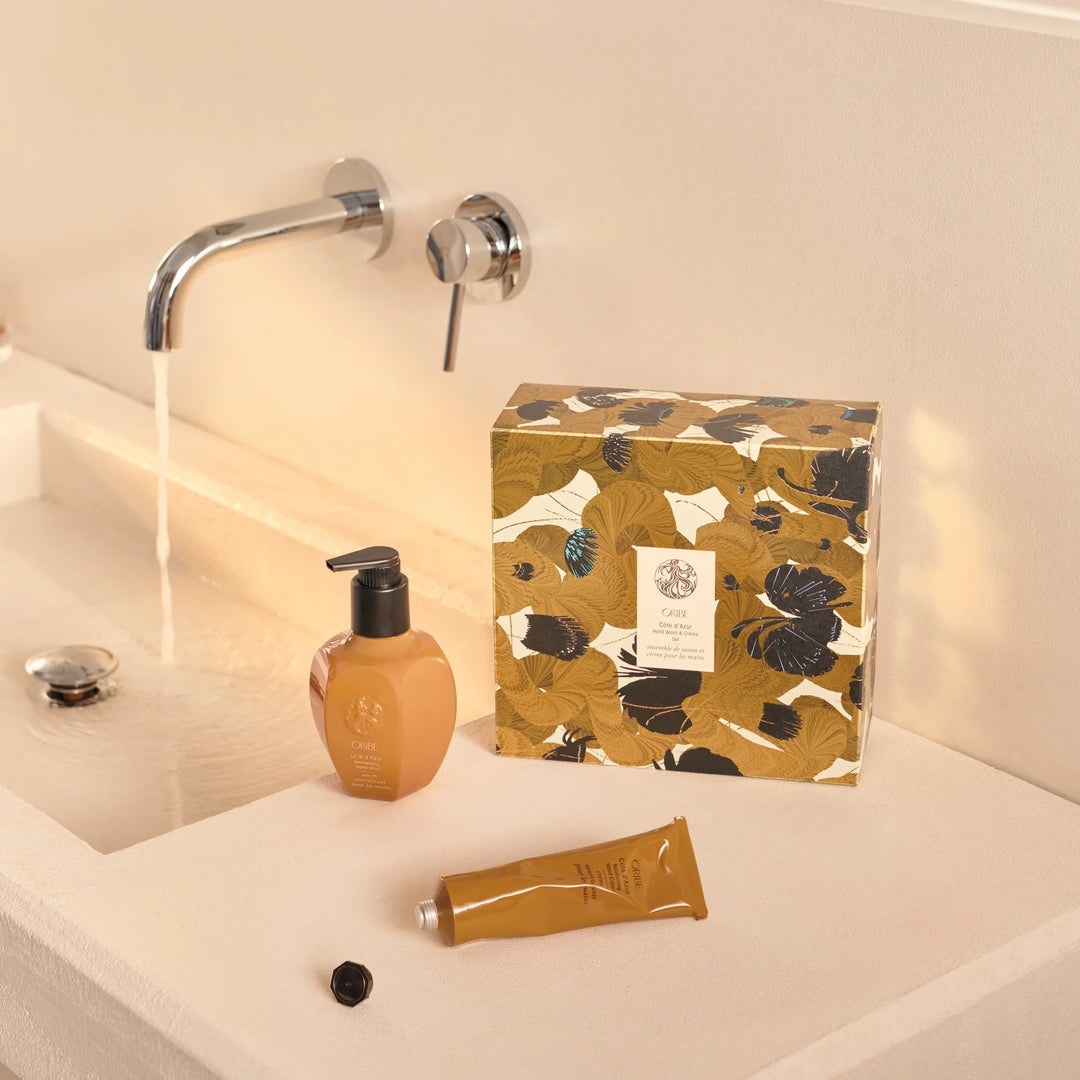 Duo mains Oribe Côte d’Azur placé sur un lavabo beige avec eau courante, accompagné du coffret fleuri doré en arrière-plan.