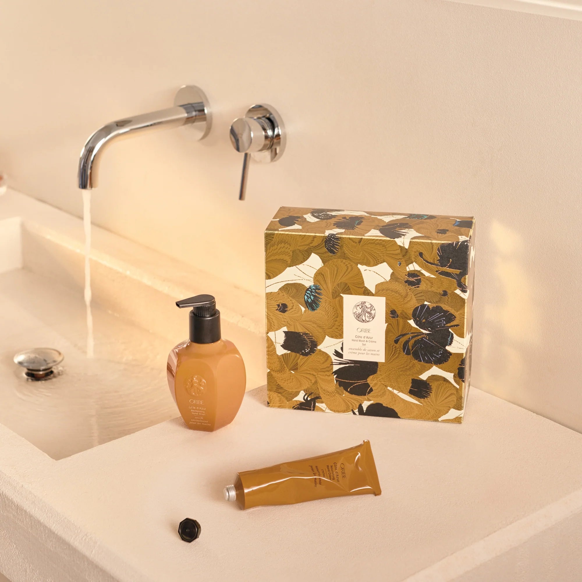 Duo mains Oribe Côte d’Azur placé sur un lavabo beige avec eau courante, accompagné du coffret fleuri doré en arrière-plan.