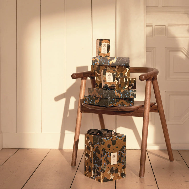 Empilement de coffrets cadeaux Oribe Côte d’Azur aux motifs floraux dorés, noirs et bleus, posés sur une chaise en bois dans un intérieur chaleureux.