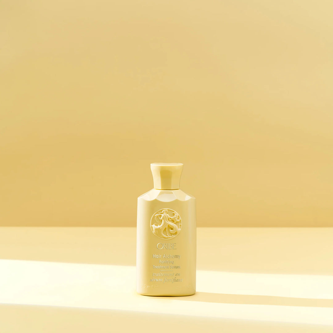 Flacon crème du sérum fortifiant Oribe Hair Alchemy, debout sur un fond beige ensoleillé, symbole d’élégance et de soin capillaire luxueux.