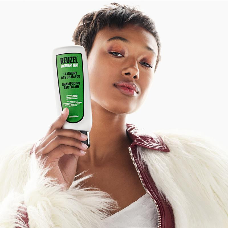 Femme aux cheveux courts tenant en main un tube de shampoing sec Reuzel Flashdry, vêtue d’un manteau en fausse fourrure blanche et d’une veste bordeaux.