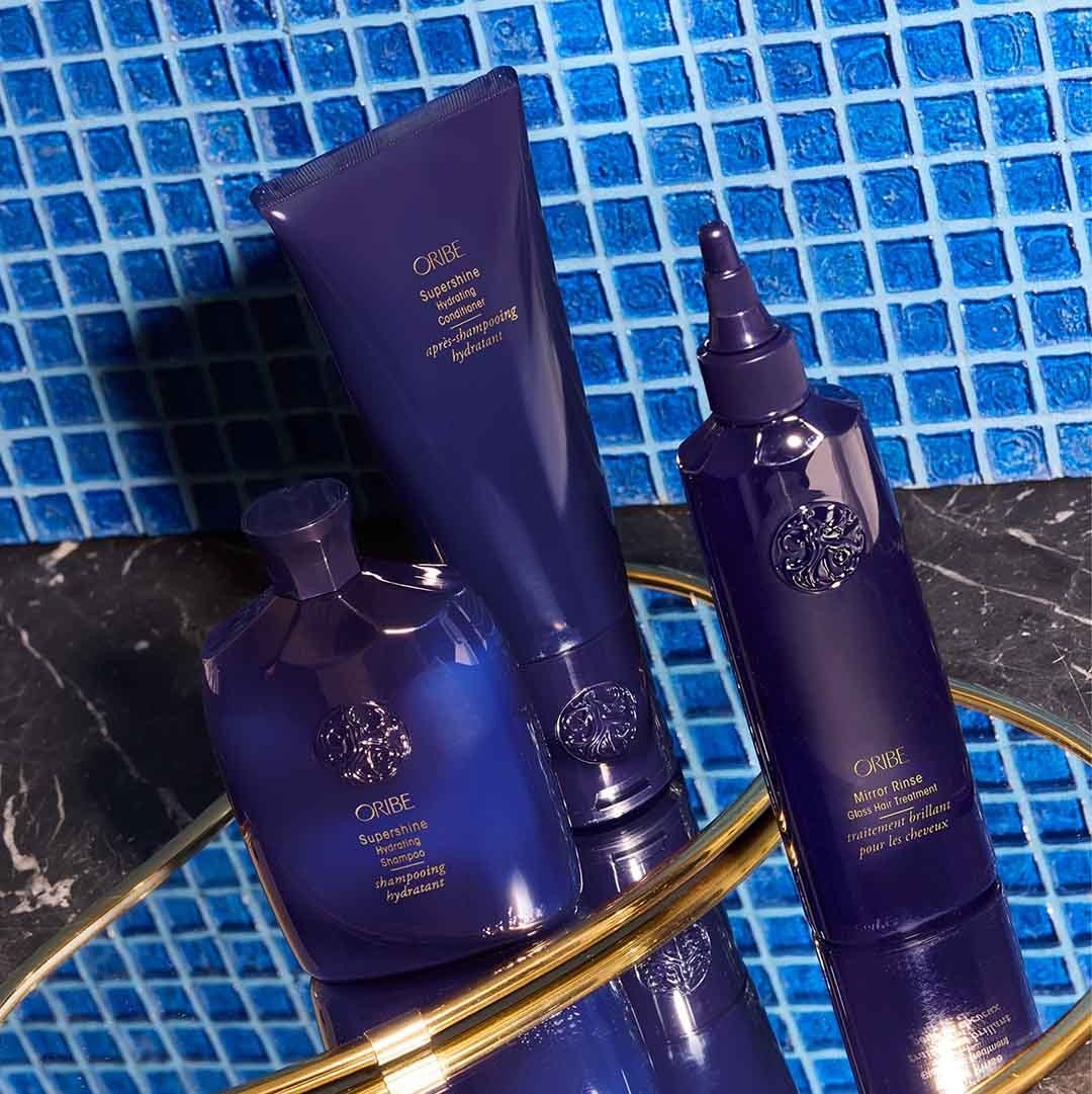 Trio de produits ORIBE (shampoing, après-shampoing, traitement) posés sur plateau doré devant un mur de mosaïque bleue, ambiance salle de bain haut de gamme.