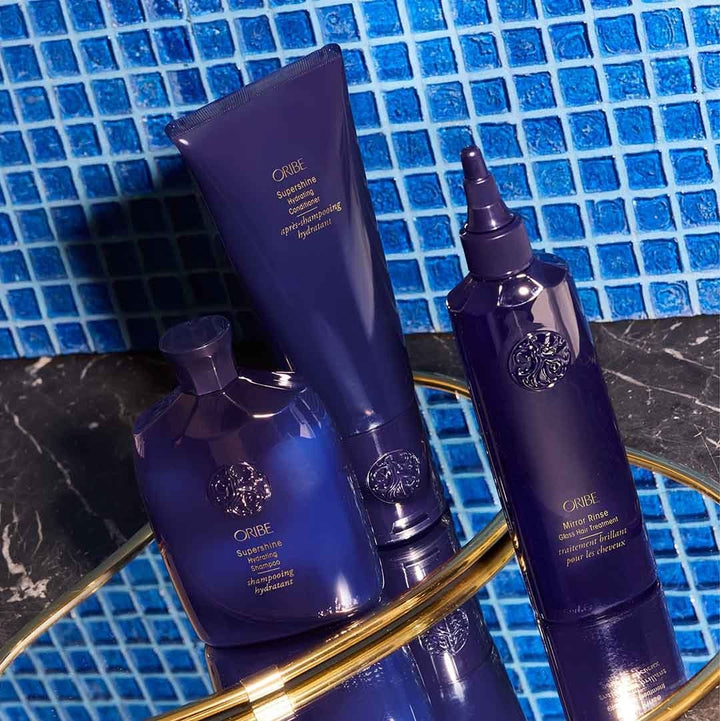 Trio de produits ORIBE (shampoing, après-shampoing, traitement) posés sur plateau doré devant un mur de mosaïque bleue, ambiance salle de bain haut de gamme.