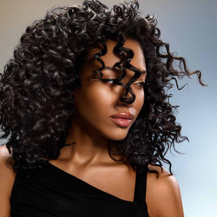 Femme aux cheveux bouclés noirs brillants, coiffure volumineuse et définie, mise en valeur du résultat après usage de soins capillaires ORIBE, fond lumineux dégradé.