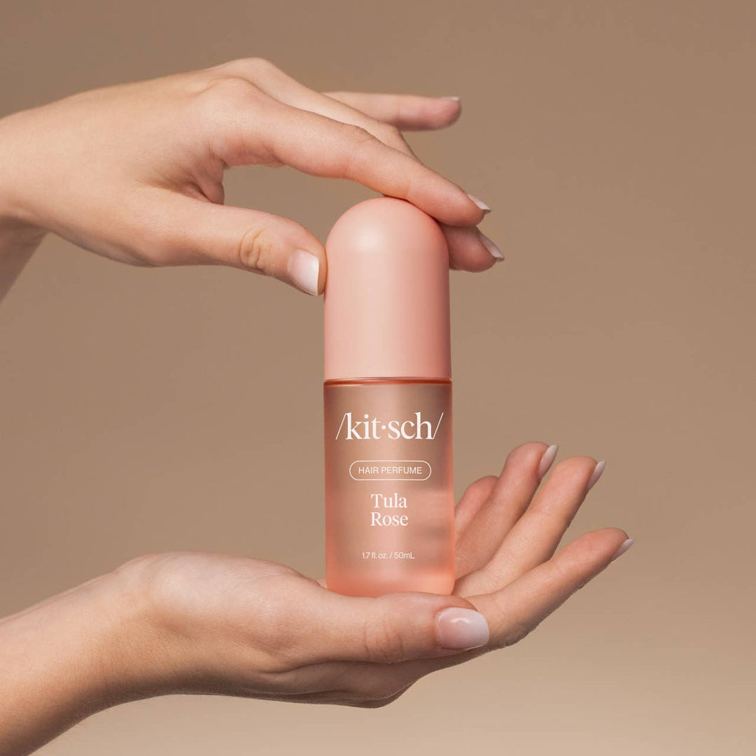 Parfum pour cheveux KITSCH Tula Rose 50 ml tenu entre deux mains, flacon rose givré avec capuchon rose mat. Parfum capillaire floral à la rose, freesia et vanille sur fond beige.
