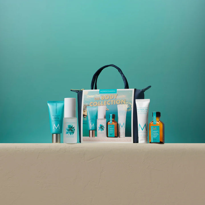 Produits Moroccanoil The Body Collection comprenant une crème pour les mains, une brume parfumée pour les cheveux et le corps, une huile Moroccanoil et un gel douche présentés devant une trousse Moroccanoil bleu marin sur fond turquoise.