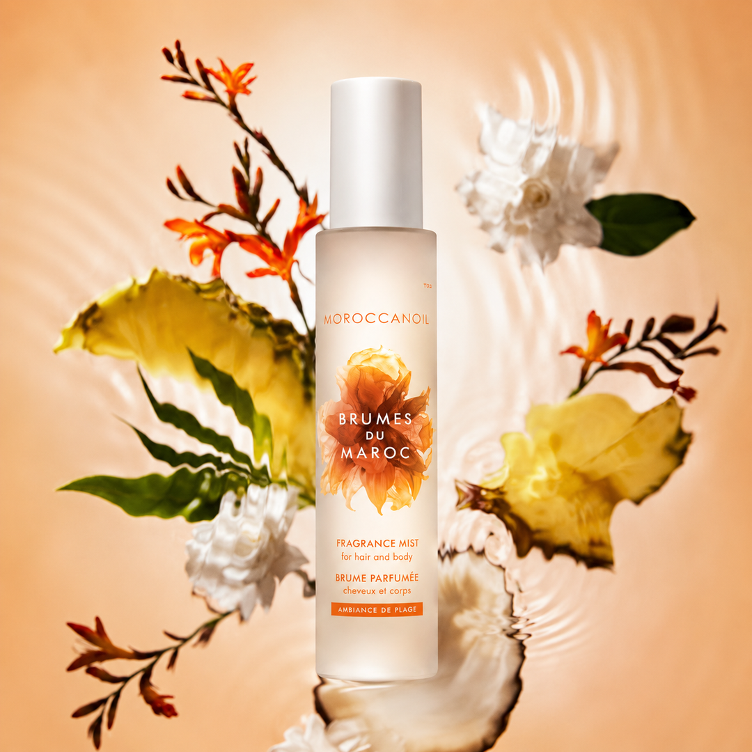 Flacon grand format Moroccanoil Brumes du Maroc Ambiance de Plage entouré de fleurs blanches tropicales et feuillage exotique sur fond beige lumineux effet aquatique.
