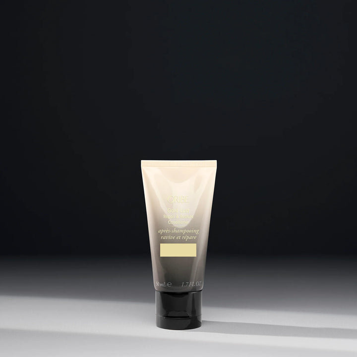Après-shampoing réparateur ORIBE Gold Lust Repair & Restore 50 ml format voyage, tube dégradé ivoire et gris foncé avec inscriptions dorées et bouchon noir, sur fond sombre.