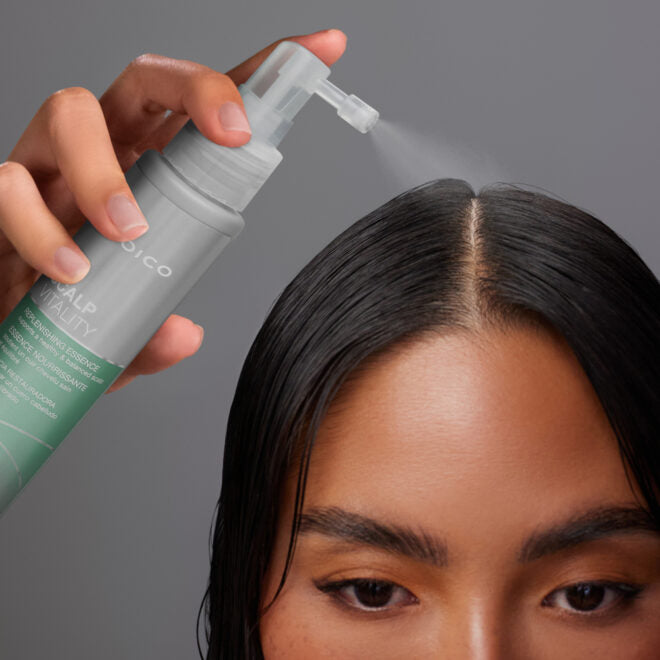 Gros plan sur une femme aux cheveux foncés et lisses appliquant l’essence nourrissante Joico Scalp Vitality directement sur la raie centrale de son cuir chevelu à l’aide du spray diffuseur, sur fond gris neutre.