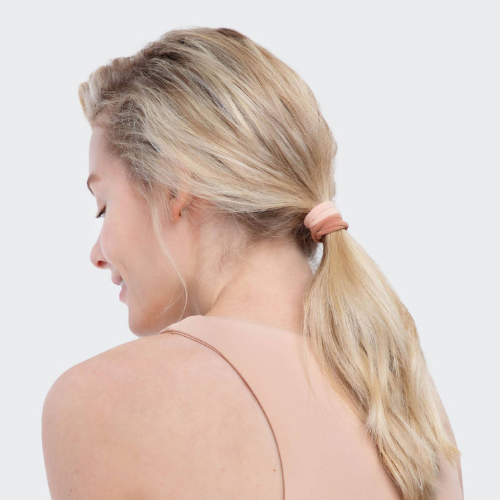 Femme portant un élastique KITSCH rose poudré dans une queue de cheval basse, accessoire cheveux écologique et sans couture sur fond beige.