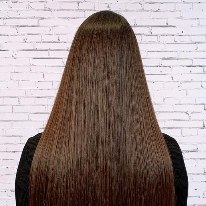 Vue arrière d’une chevelure longue, lisse et brillante de couleur châtain foncé, parfaitement coiffée, devant un mur de briques blanches. Les cheveux apparaissent doux, disciplinés et sans frisottis, résultat d’un lissage thermique.