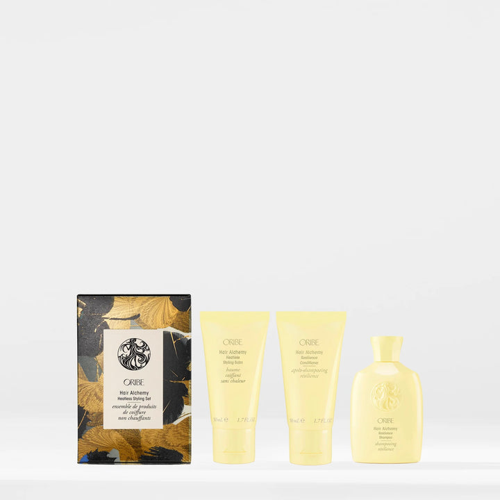 Coffret Oribe Hair Alchemy Heatless Styling Set sur fond blanc, avec trois produits posés à côté : baume coiffant, après-shampooing et shampooing résilience en flacons jaune pastel, au design raffiné.