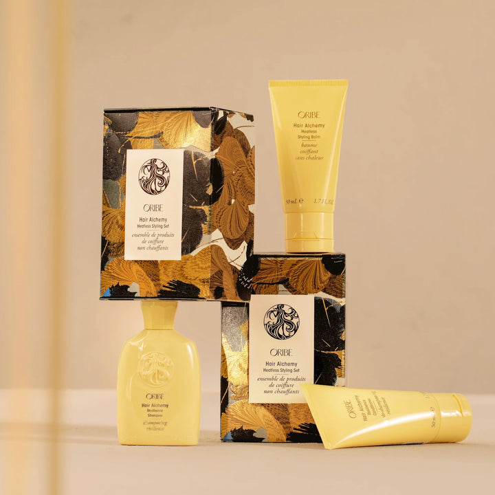 Coffret Oribe Hair Alchemy Heatless Styling Set présenté sur fond beige, avec trois produits capillaires jaunes : shampooing, après-shampooing et baume coiffant sans chaleur. Boîte décorative noire et dorée au motif botanique élégant.