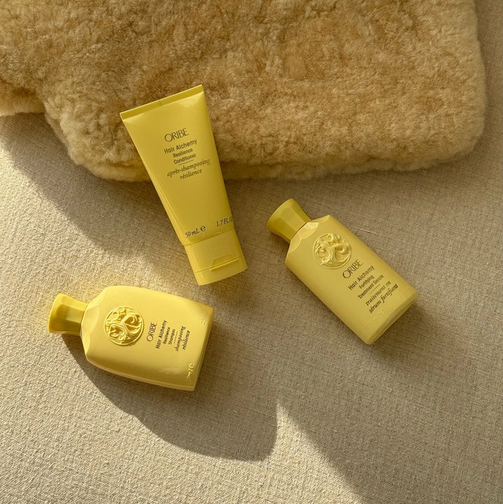 Produits capillaires Oribe Hair Alchemy en format voyage, posés sur une surface beige texturée avec une couverture en fausse fourrure. Trois flacons jaunes : shampooing, après-shampooing et traitement fortifiant. Ambiance douce et cocooning.