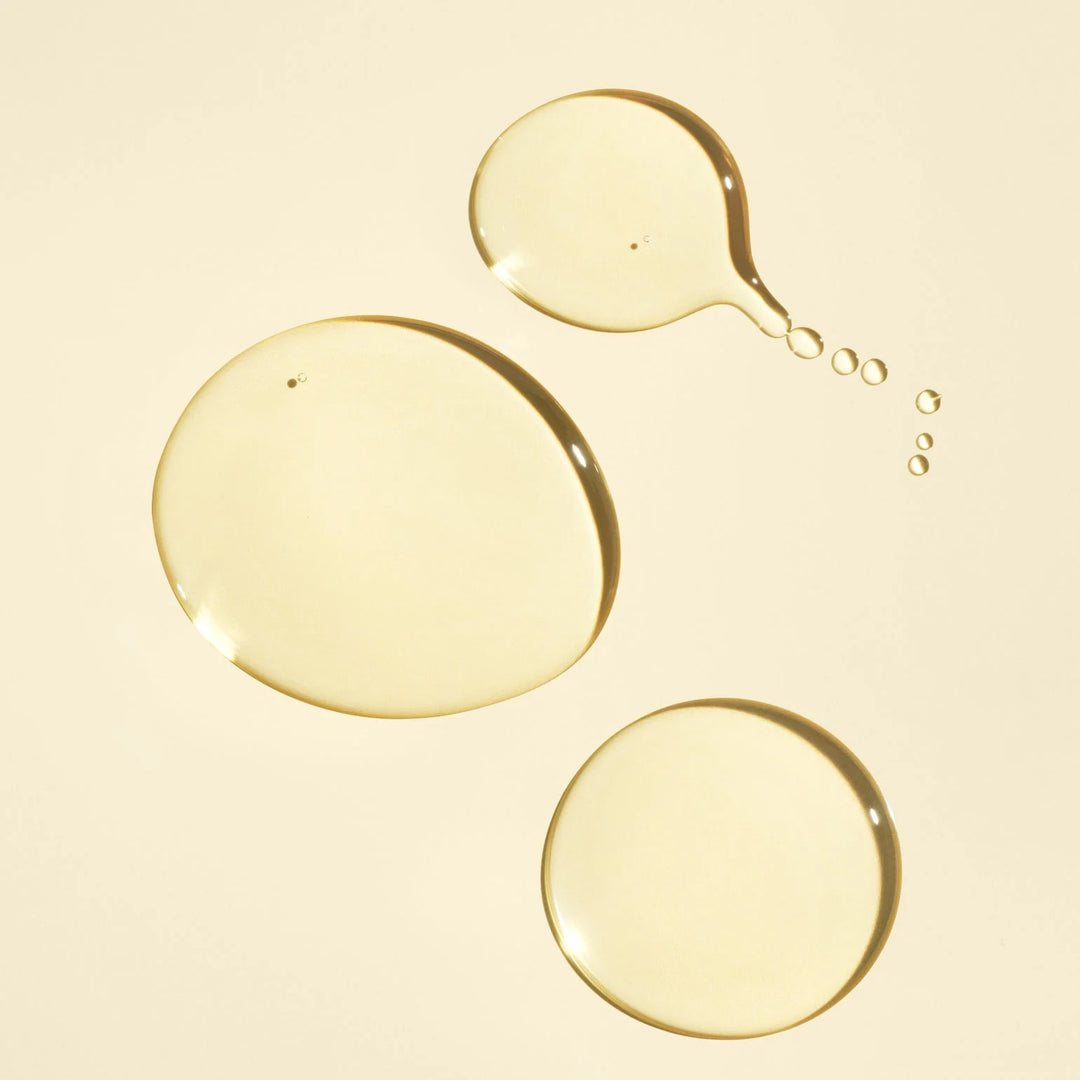Texture dorée et translucide de l'huile nourrissante ORIBE Gold Lust, présentée en gouttes brillantes sur un fond beige clair.